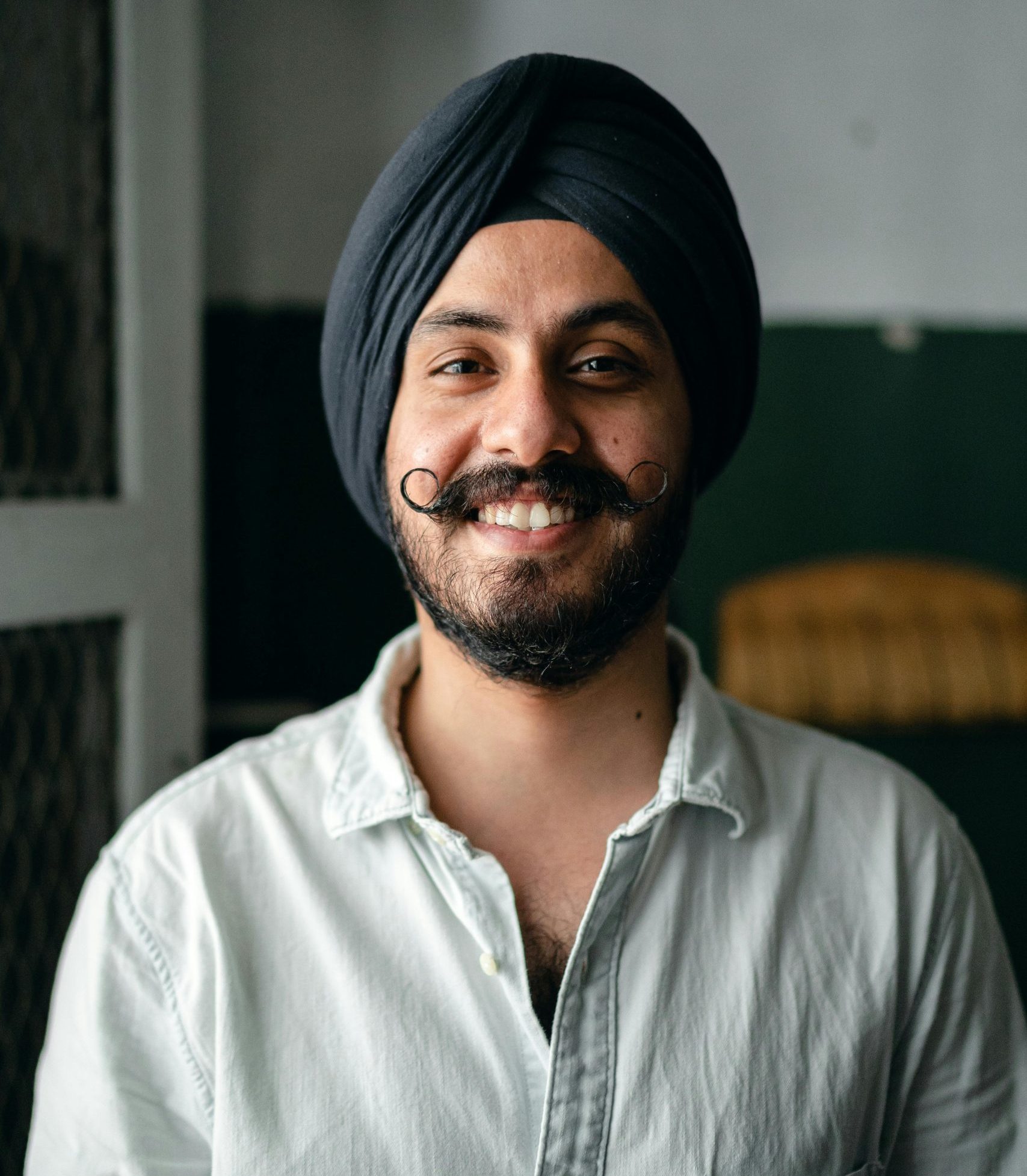 Positive bearded Indian male in shirt and turban standing in room and smiling while looking at camera